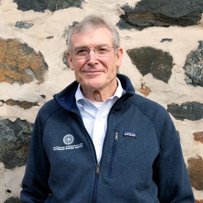The image shows an older man with glasses, wearing a blue jacket over a white shirt. He is standing in front of a stone wall. The man has short, gray hair and appears to be smiling slightly. The jacket has a logo on the left side. The background is a textured stone wall.